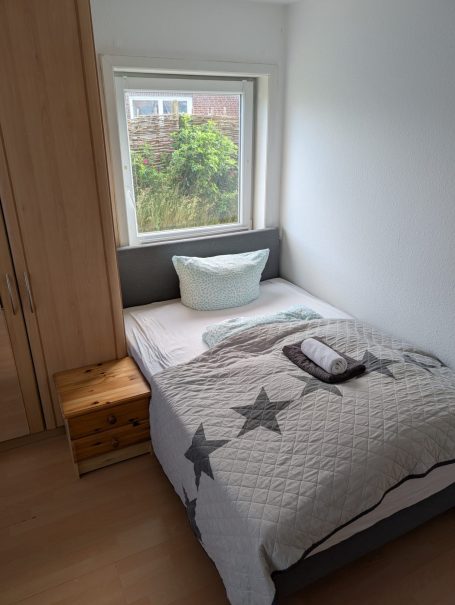 Blick auf ein Schlafzimmer mit Bett, Kissen, Decke und Fenster zur grünen Landschaft.